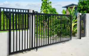 Black metal sliding gate at a modern residential entrance, bordered by concrete posts and surrounded by greenery in a landscaped outdoor setting.